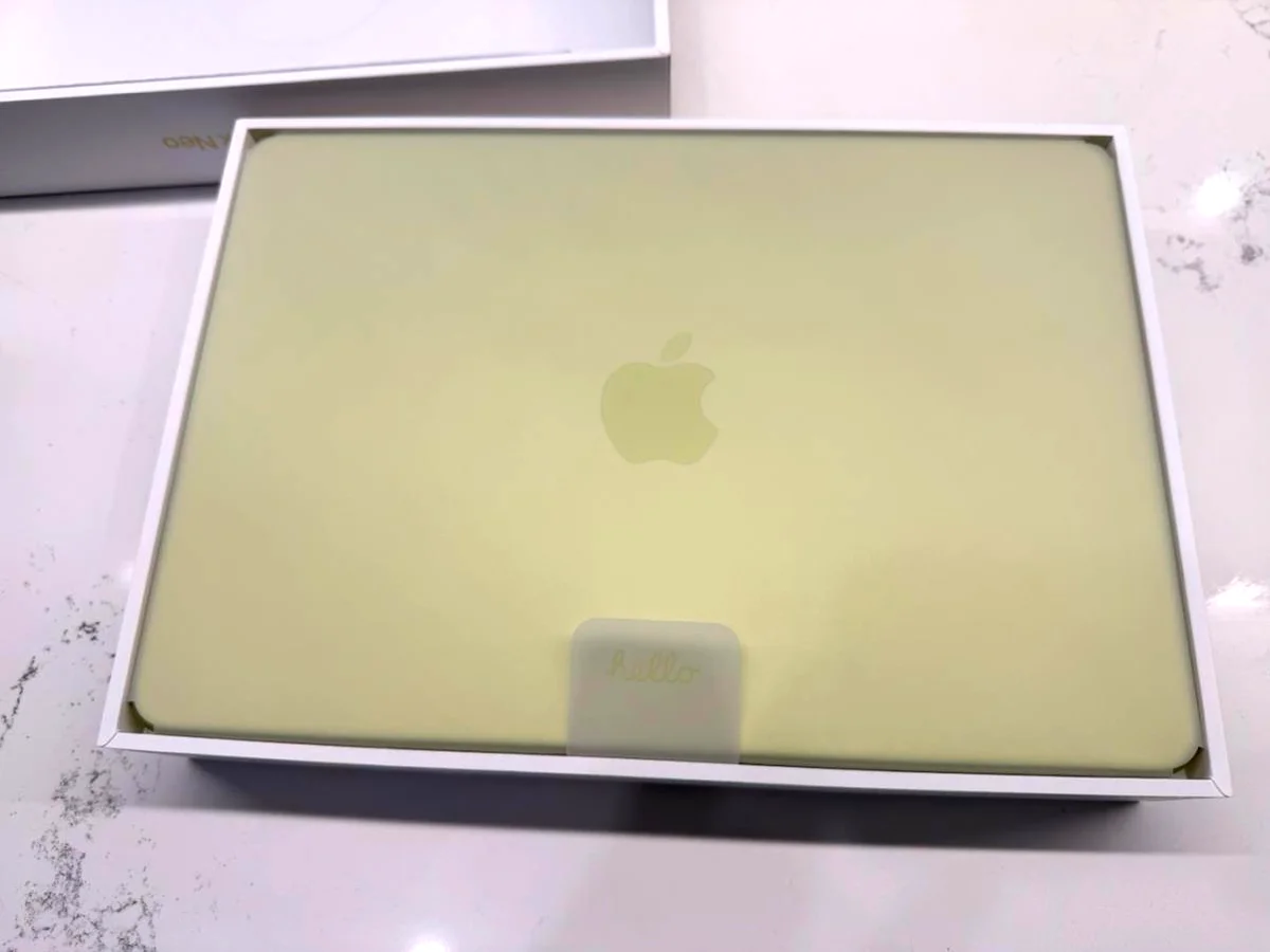 MacBook Neo in citrus colour, lid closed inside the open Apple box with 'hello' tag visible
